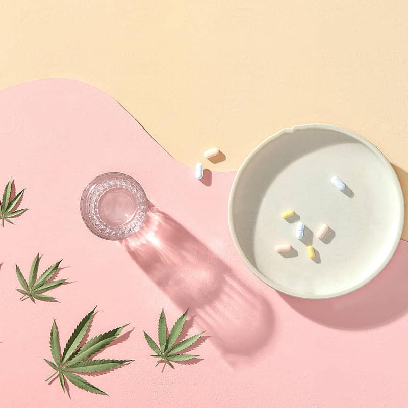 glass of water and mental health pills are pictured next to a few small cannabis leaves
